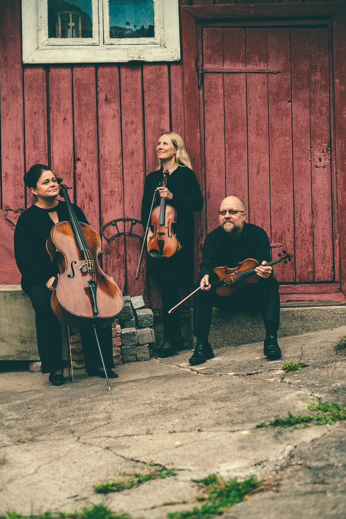 En kvinne med en cello, en kvinne med en bratsj og en mann med en fiolin. I bakgrunnen er en rød låvevegg.