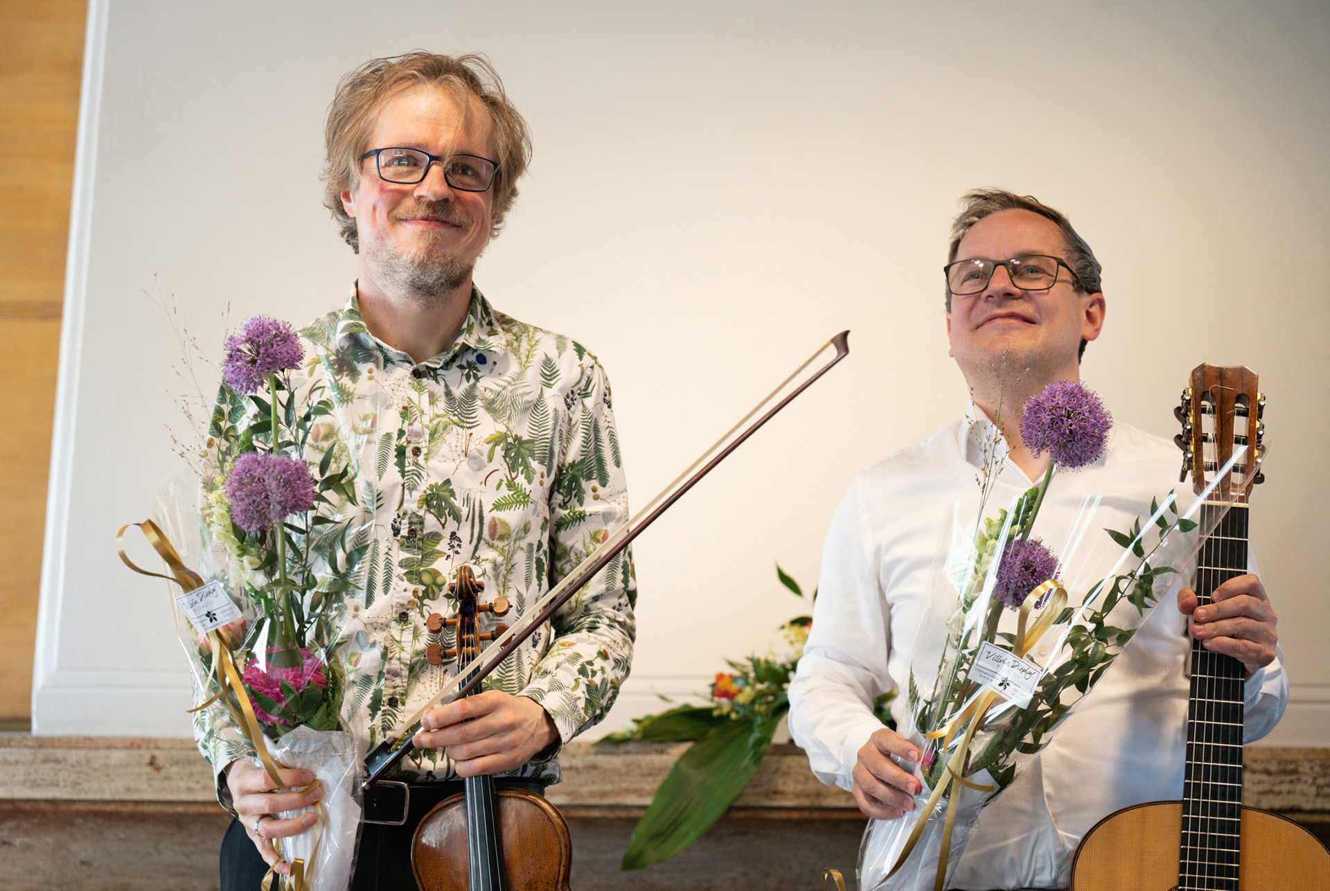 To menn med blomsterbuketter i hendene. Den ene holder en fiolin, den andre en gitar.