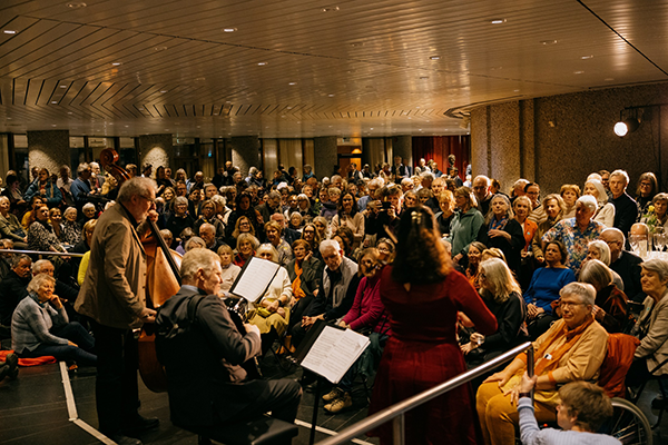 Musikere med cello, fiolin og trekkspill foran et fullsatt publikum