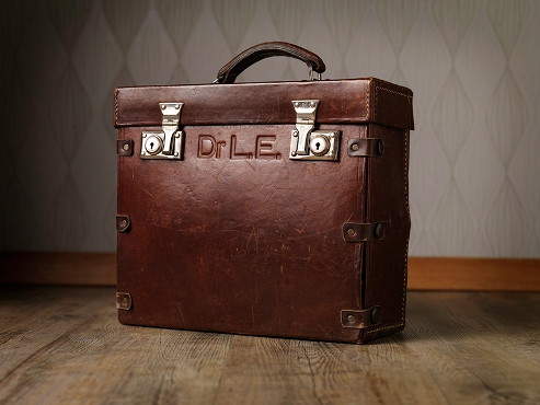 En  brun koffert plassert i et rom med tregulv. Står Dr. L.E inngravert på den.