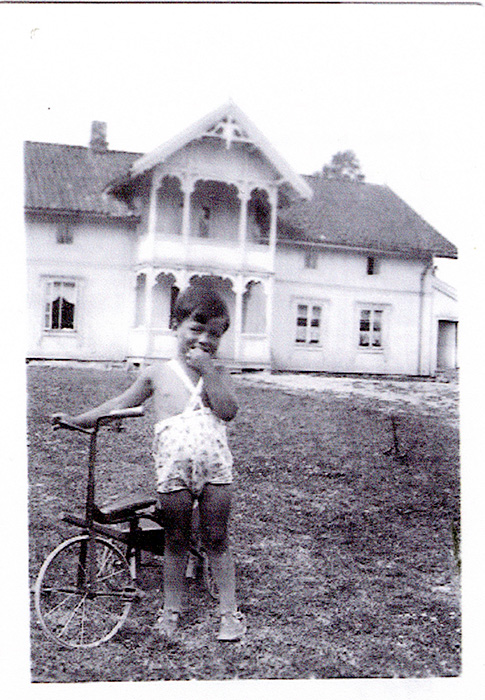 Liten gutt i kortbukser ved siten av en sykkel. Stort, hvitt hus i bakgrunnen