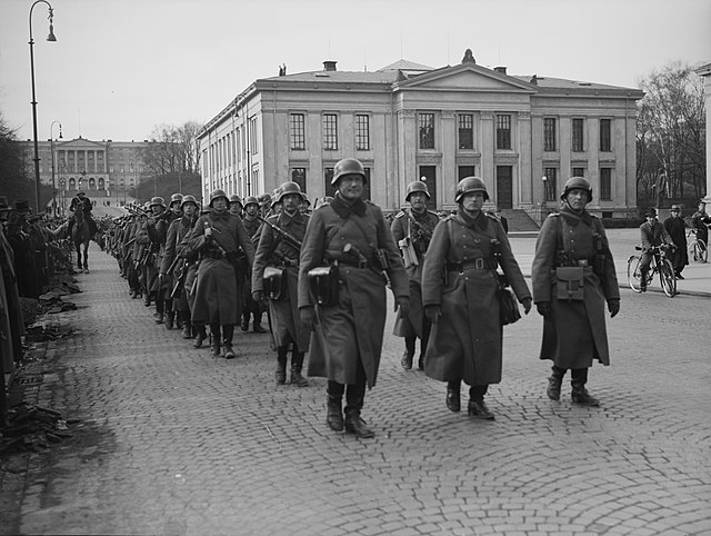 Soldater som marsjerer tre i rekken. I bakgrunnen skimtes en stor, hvit murbygning