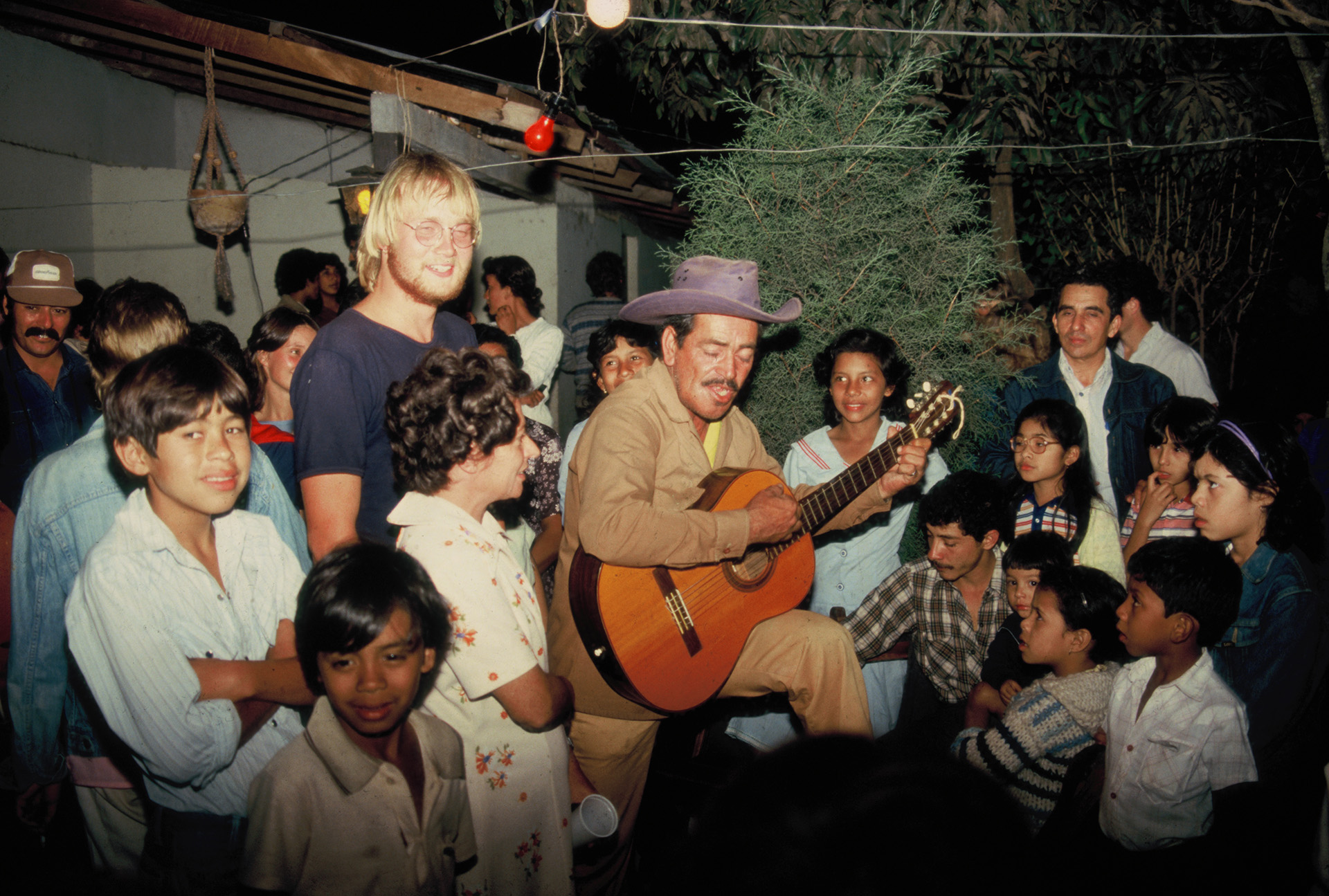 Gruppe mennesker sitter sammen i en ring rundt en person med cowboyhatt som spiller gitar. Fargerike klær.