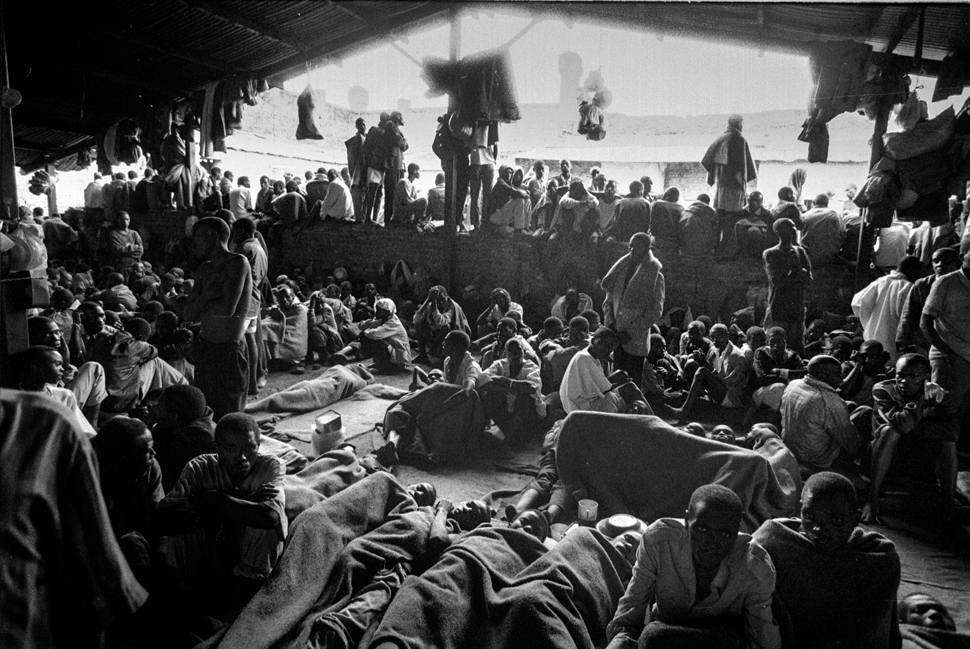 Stor gruppe mennesker som delvis ligger på bakken under tepper og delvis sitter eller står. Over dem er det et slags tak. Bildet er i sort-hvitt.